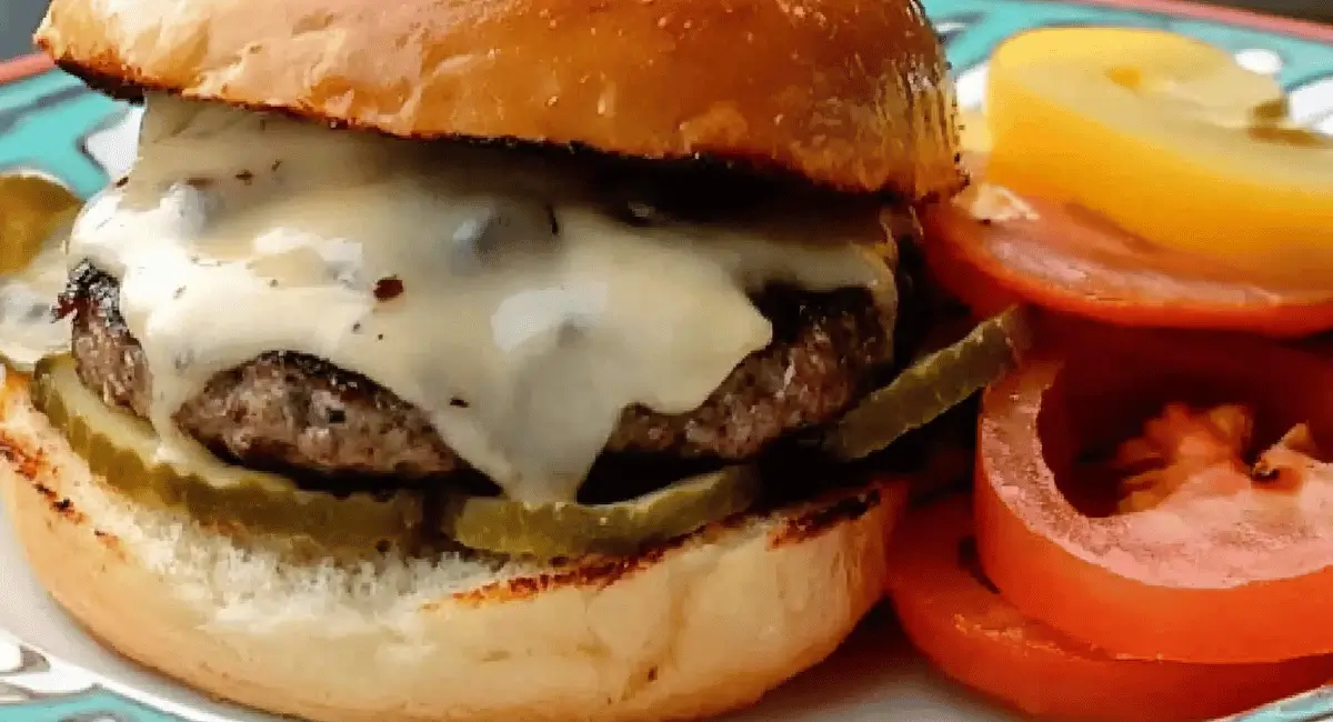 A cooked burger with melted white cheese, pickles, and tomatoes on a plate.