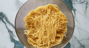 Overhead view of cooked spaghetti noodles piled in a metal mesh strainer resting on a white marble surface.