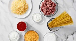 Overhead view of various ingredients arranged on a white marble countertop, including uncooked spaghetti, ground meat, shredded cheese (two types), tomato sauce, and ricotta cheese in separate bowls.