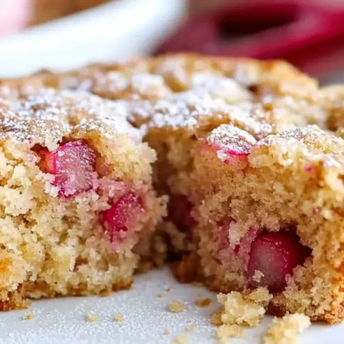 A round rhubarb cake with a wedge missing, revealing the juicy rhubarb chunks inside, generously sprinkled with powdered sugar and served on a white plate.