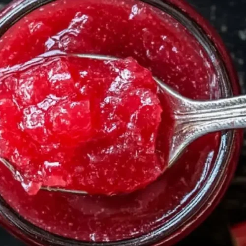 A silver spoon scoops a generous portion of bright pink rhubarb jam from an open glass jar, with pieces of raw rhubarb scattered on a dark surface.