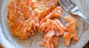 Flaked cooked salmon on a light brown plate with a fork, showing a key ingredient prepared for a salmon cake recipe.