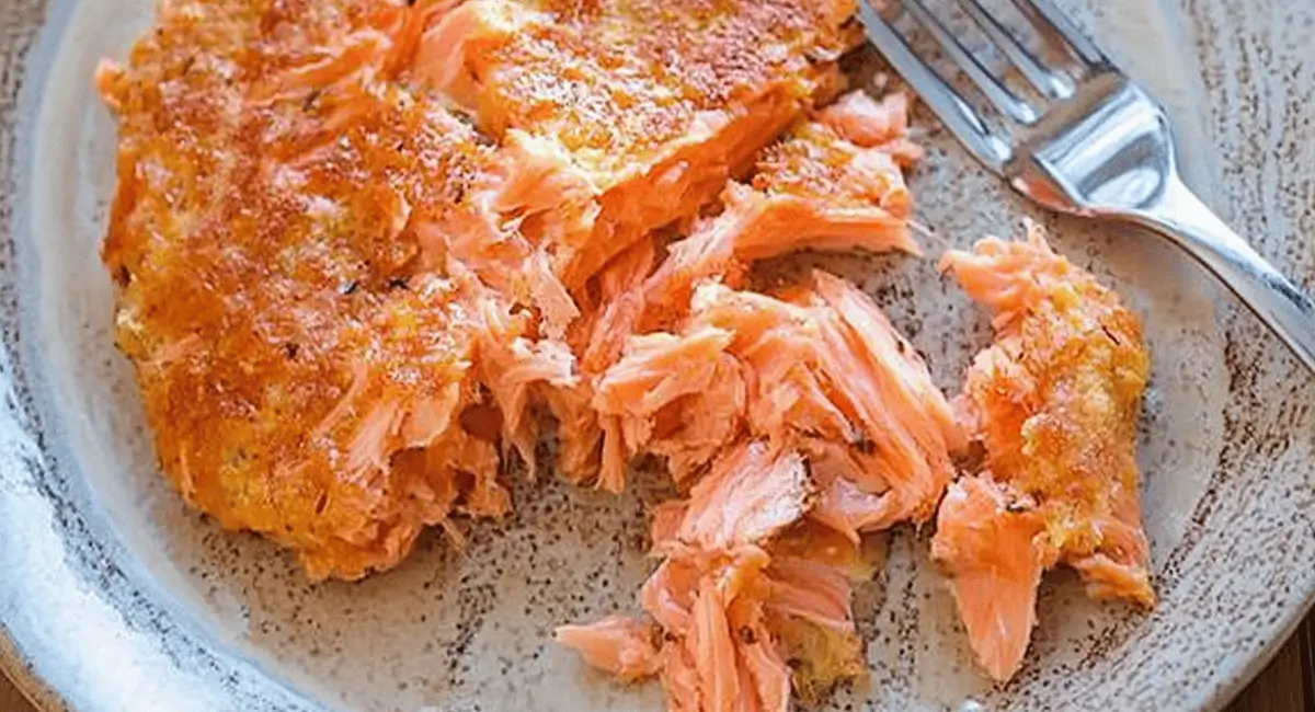 Flaked cooked salmon on a light brown plate with a fork, showing a key ingredient prepared for a salmon cake recipe.