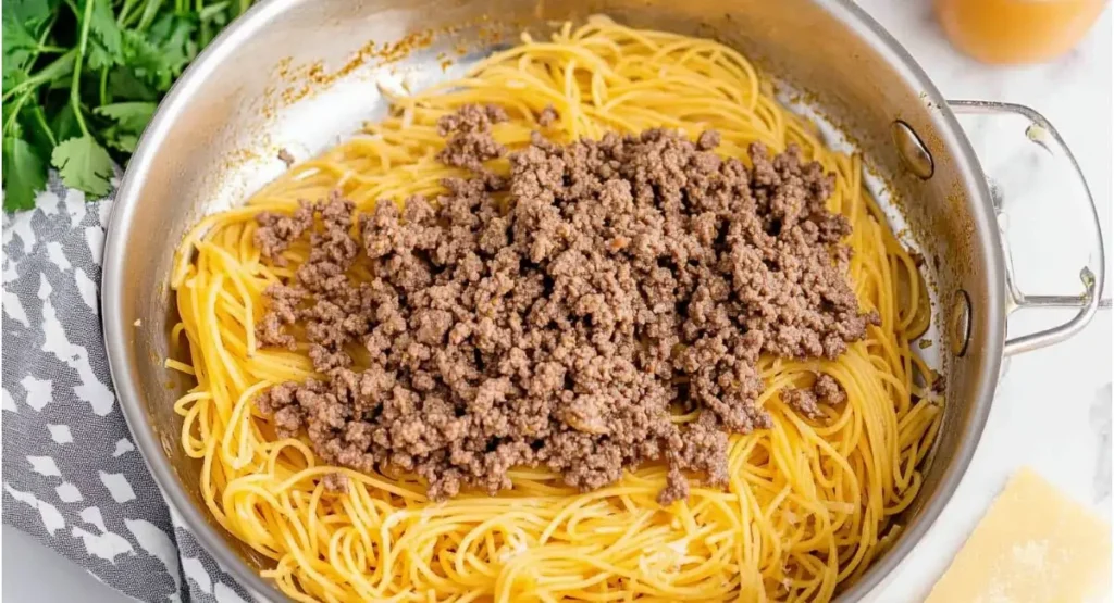 A stainless steel pan containing cooked spaghetti topped with browned ground beef, the beginnings of taco spaghetti.