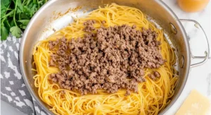 A stainless steel pan containing cooked spaghetti topped with browned ground beef, the beginnings of taco spaghetti.