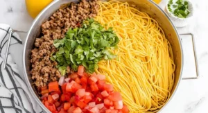 A stainless steel pan with distinct sections of cooked spaghetti, browned ground beef, chopped tomatoes and onions, and fresh cilantro, preparing for taco spaghetti.