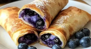 Three Air Fryer Blueberry Chimichangas stacked on a white plate, dusted with powdered sugar and surrounded by fresh blueberries.