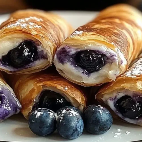 Close-up of several golden brown Air Fryer Blueberry Chimichangas on a white plate, dusted with powdered sugar, with fresh blueberries scattered at the base.