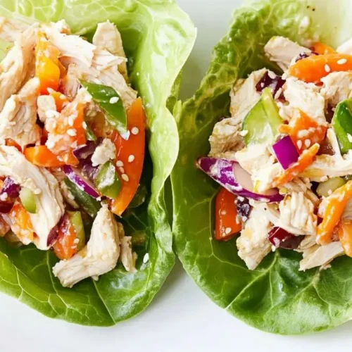 Two Asian Chicken Salad Lettuce Wraps on a white plate, filled with shredded chicken, julienned carrots, diced red and green vegetables, and topped with sesame seeds.