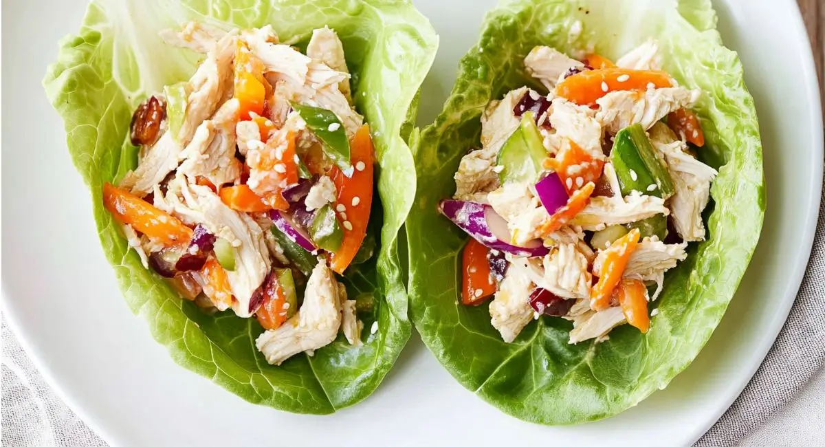 Two Asian Chicken Salad Lettuce Wraps on a white plate, filled with shredded chicken, julienned carrots, diced red and green vegetables, and topped with sesame seeds.
