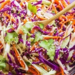 A clear glass bowl filled with Asian Slaw with Sesame Dressing and a wooden spoon.