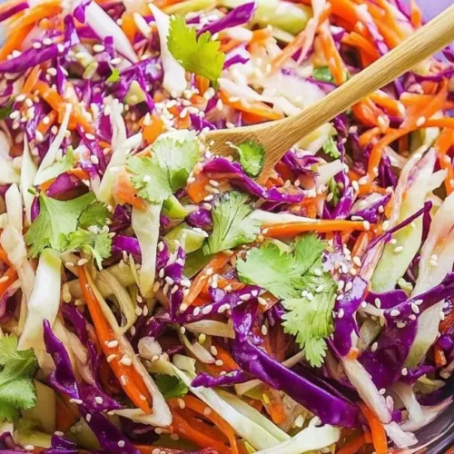 A clear glass bowl filled with Asian Slaw with Sesame Dressing and a wooden spoon.