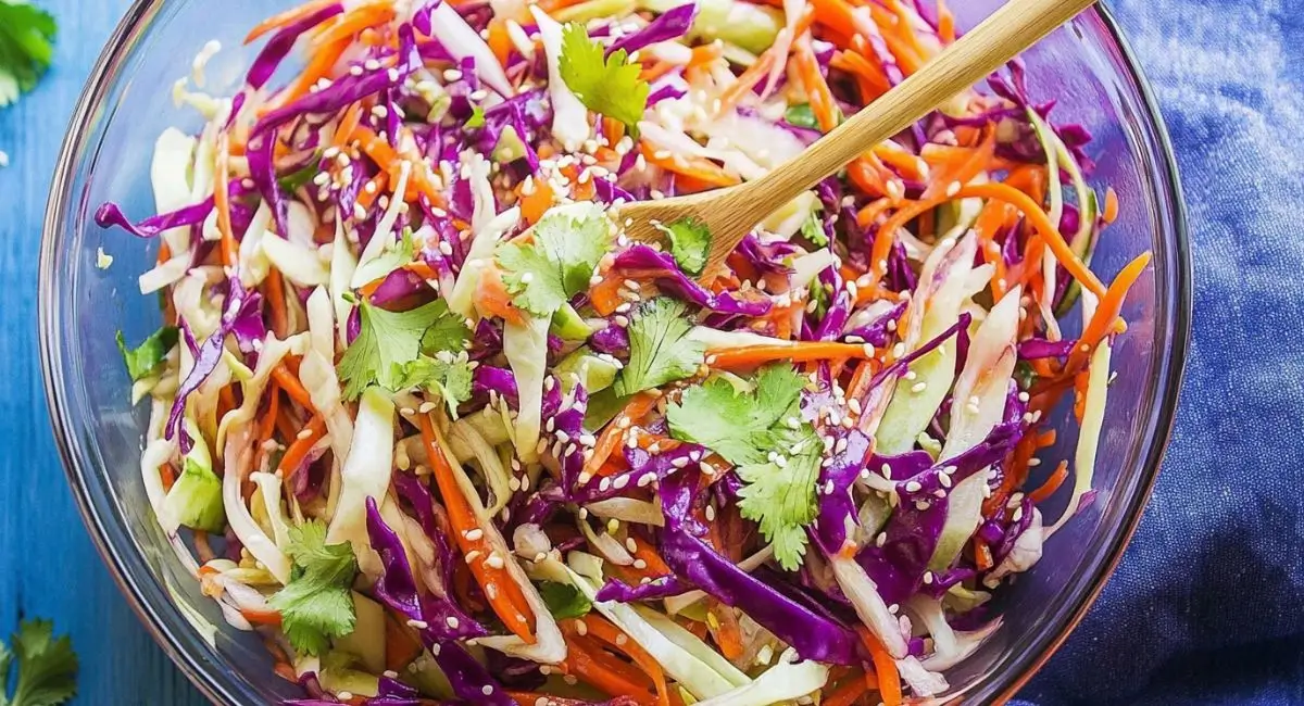 A clear glass bowl filled with Asian Slaw with Sesame Dressing and a wooden spoon.