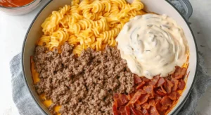 An overhead view of the uncooked components of a Bacon Cheeseburger Casserole arranged in a gray skillet: cooked spiral pasta, a creamy sauce, seasoned ground beef, and crispy bacon pieces.