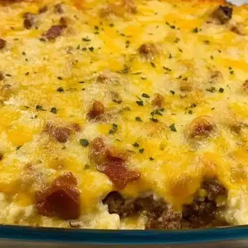 A close-up of a partially eaten Bacon Cheeseburger Casserole in a clear glass baking dish, showing layers of creamy sauce, ground beef, bacon pieces, and melted cheddar cheese topped with fresh parsley.