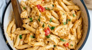 An overhead shot shows a white bowl filled with creamy Cajun Cream Cheese Chicken Pasta. Penne noodles, tender chicken chunks, and diced red tomatoes are coated in a rich sauce and garnished with fresh parsley. A wooden spoon rests in the pasta.