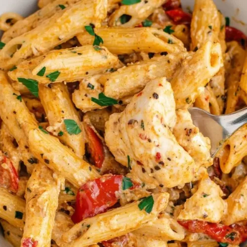 A close-up overhead shot shows a creamy bowl of Cajun Cream Cheese Chicken Pasta with penne noodles, tender chicken pieces, diced tomatoes, and fresh parsley, being lifted with a silver fork with a red handle.
