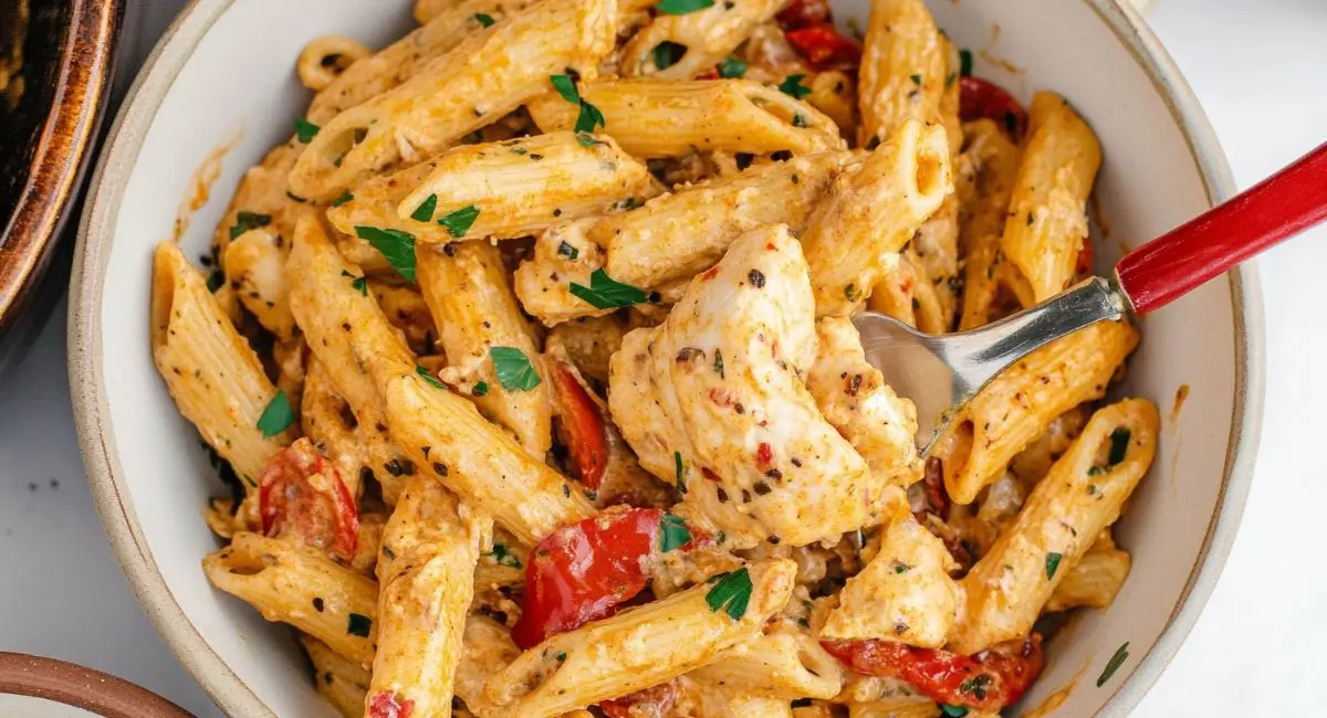 A close-up overhead shot shows a creamy bowl of Cajun Cream Cheese Chicken Pasta with penne noodles, tender chicken pieces, diced tomatoes, and fresh parsley, being lifted with a silver fork with a red handle.