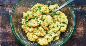 A clear glass bowl containing a mixture of shredded cauliflower and other ingredients, being stirred with a metal spoon, for making Cauliflower Fritters.