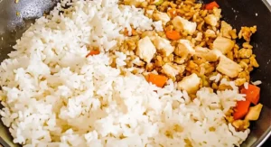 A pan containing a partially mixed Chicken Fried Rice Recipe, with distinct sections of white rice and a mixture of seasoned rice with chicken and vegetables.