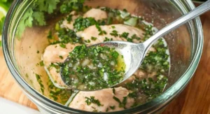 A silver spoon lifts a generous spoonful of vibrant green and yellow Cilantro Lime Chicken marinade from a clear glass bowl containing raw chicken breasts.