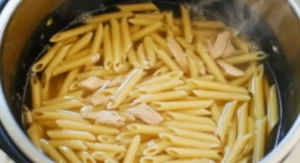 An overhead view of penne pasta cooking in a clear broth within an Instant Pot, with visible pieces of shredded chicken scattered among the pasta. This is a step in making Creamy Buffalo Garlic Parmesan Pasta.