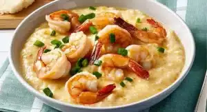 A white bowl filled with creamy Shrimp and Grits, adorned with cooked shrimp and chopped green onions, sits on a blue patterned surface.