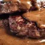 A close-up shot of two juicy Hamburger Steaks with Brown Gravy stacked on a white plate, with extra gravy pooled around the base.