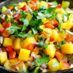 A dark bowl filled with a mixed Indian Style Chaat Masala Mango Salad Recipe, featuring diced mangoes, tomatoes, red onions, cucumbers, and cilantro, tossed together.