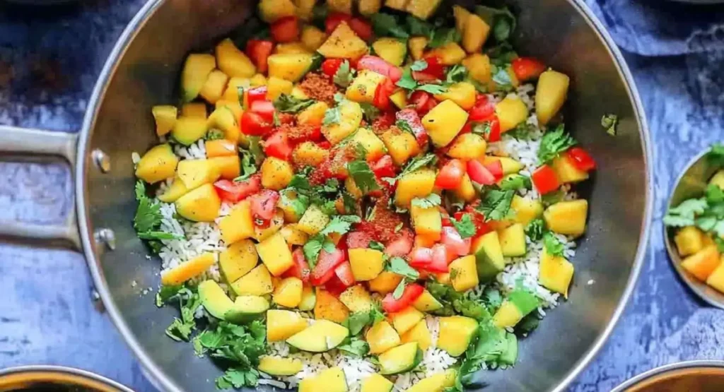 A stainless steel pan filled with ingredients for Indian Style Chaat Masala Mango Salad Recipe, including diced mangoes, tomatoes, cilantro, spices, and possibly avocado, over a base of white rice.