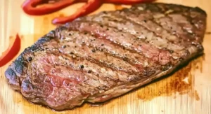Grilled juicy flank steak with visible cross marks on a wooden board with roasted red pepper. Juicy Flank Steak Recipe.