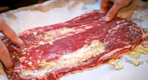Raw flank steak being seasoned with dry spices on parchment paper, hands visible. Juicy Flank Steak Recipe.