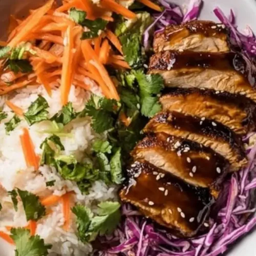 A white bowl filled with a colorful Korean Chicken Lunch Meal Prep. It contains white rice topped with shredded carrots and cilantro on one side, and thinly sliced, glazed chicken breast sprinkled with sesame seeds and cilantro on a bed of shredded purple cabbage on the other side.