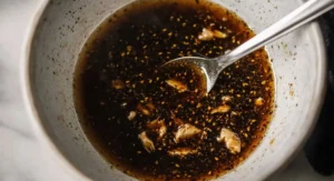 A light grey speckled bowl containing a dark brown, glossy Korean Chicken Marinade with visible sesame seeds and pieces of garlic, stirred with a silver spoon.