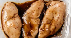 Three raw chicken breasts marinating in a clear plastic bag filled with a dark brown Korean Chicken Marinade, showing visible red pepper flakes and garlic.