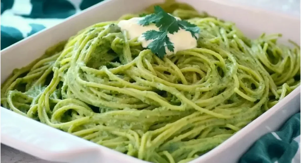 A white rectangular baking dish filled with creamy green spaghetti, garnished with a dollop of white cream and a fresh cilantro sprig. Mexican Green Spaghetti Recipe.