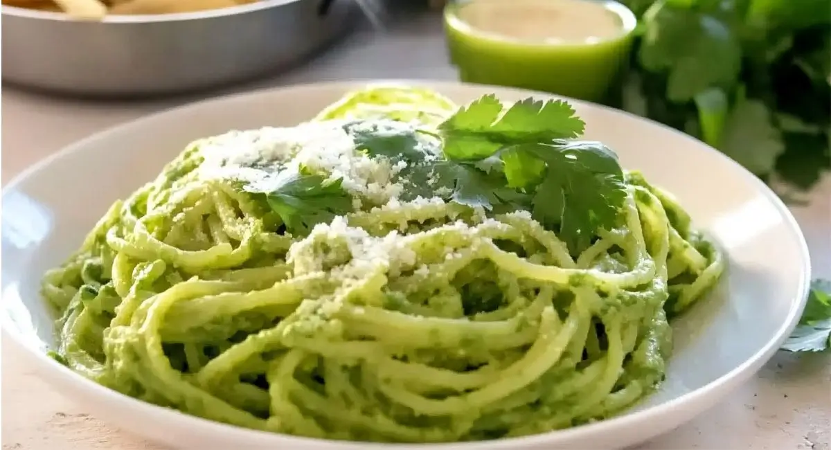 A white plate of green spaghetti topped with grated white cheese and cilantro, with a lime wedge and cilantro sprigs in the background. Mexican Green Spaghetti Recipe.