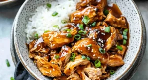 A white and grey speckled bowl filled with white rice and a generous serving of saucy Slow Cooker Honey Teriyaki Chicken, garnished with green onions and sesame seeds.