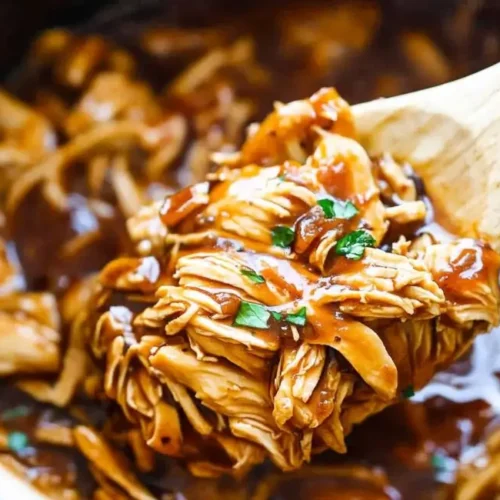 A wooden spoon lifting a large scoop of shredded, saucy Slow Cooker Honey Teriyaki Chicken from a white slow cooker, garnished with fresh herbs.