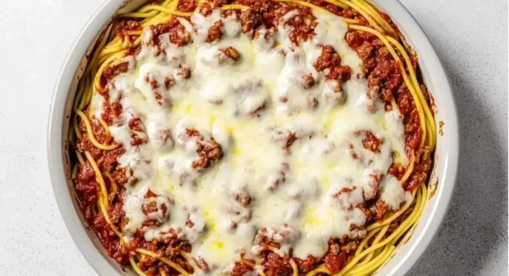Baked spaghetti pie in a round white baking dish, topped with melted cheese and meat sauce.