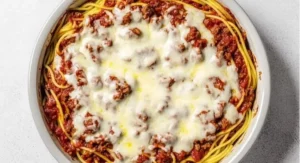 Baked spaghetti pie in a round white baking dish, topped with melted cheese and meat sauce.
