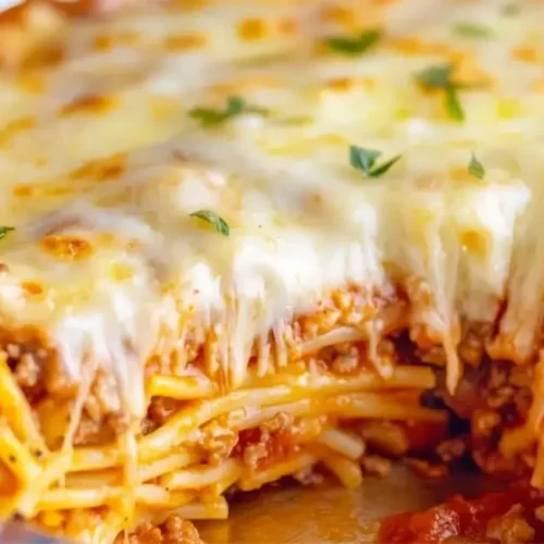 Close-up of a slice of layered Spaghetti Pie being removed from a round foil pan, showcasing the spaghetti, meat sauce, and melted cheese layers.