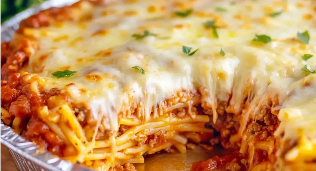 Close-up of a slice of layered Spaghetti Pie being removed from a round foil pan, showcasing the spaghetti, meat sauce, and melted cheese layers.