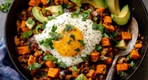 A single serving of Tex-Mex Sweet Potato Hash in a skillet, topped with a fried egg, avocado, and cilantro.