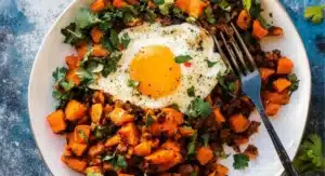 A plate of flavorful Tex-Mex Sweet Potato Hash topped with a fried egg and fresh cilantro, with a fork resting on the side.