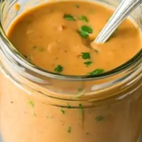 A jar filled with smooth, light brown sauce with green flecks, a metal spoon inside, set against a slightly blurred background with a hint of blue cloth, representing a component of apricot chicken recipe baked.