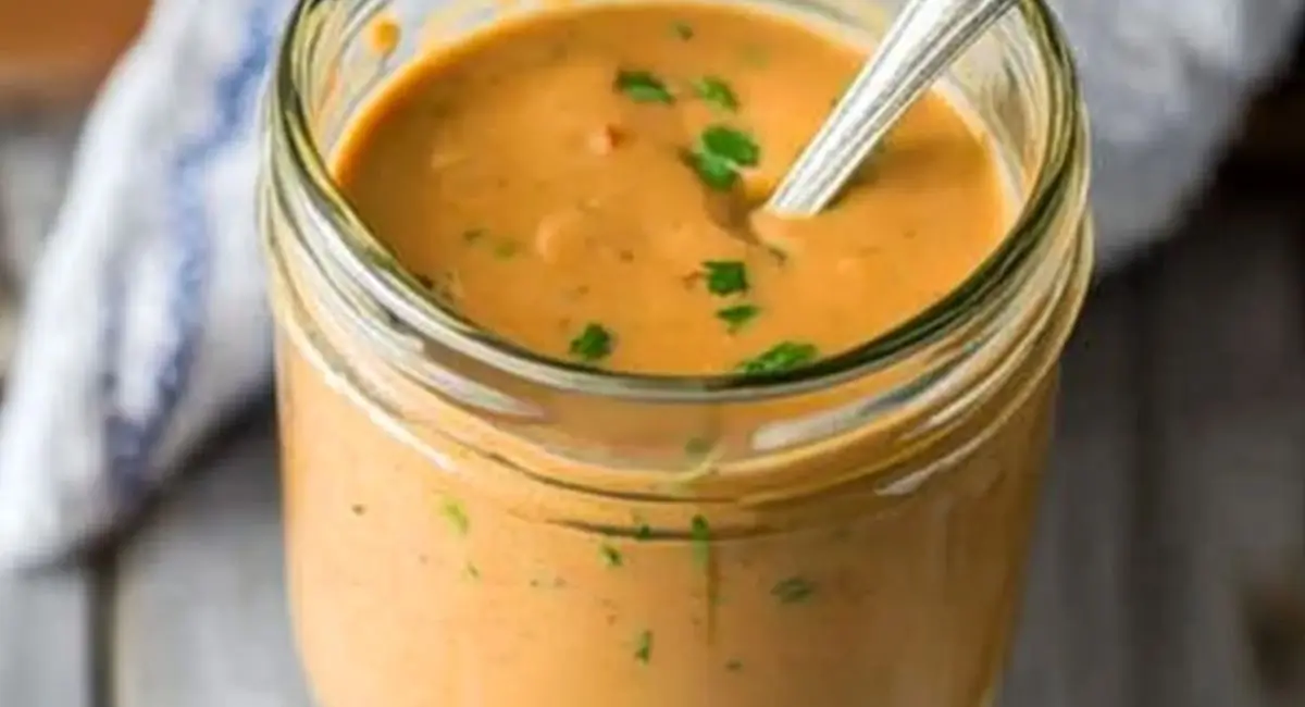 A jar filled with smooth, light brown sauce with green flecks, a metal spoon inside, set against a slightly blurred background with a hint of blue cloth, representing a component of apricot chicken recipe baked.