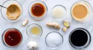 Overhead view of various ingredients in small glass bowls on a white marble surface, including peanut butter, soy sauce, rice vinegar, sugar, and ginger, preparing for an apricot chicken recipe baked.