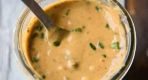 Close-up of a creamy, light brown sauce with green flecks in a clear glass jar, a metal spoon partially submerged in it. Apricot chicken recipe baked.
