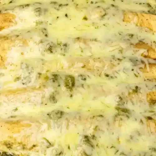 A close-up overhead shot of a white baking dish filled with The Best Green Chili Chicken Enchiladas Recipe, featuring golden-brown tortillas smothered in a creamy green chili sauce and melted white cheese.